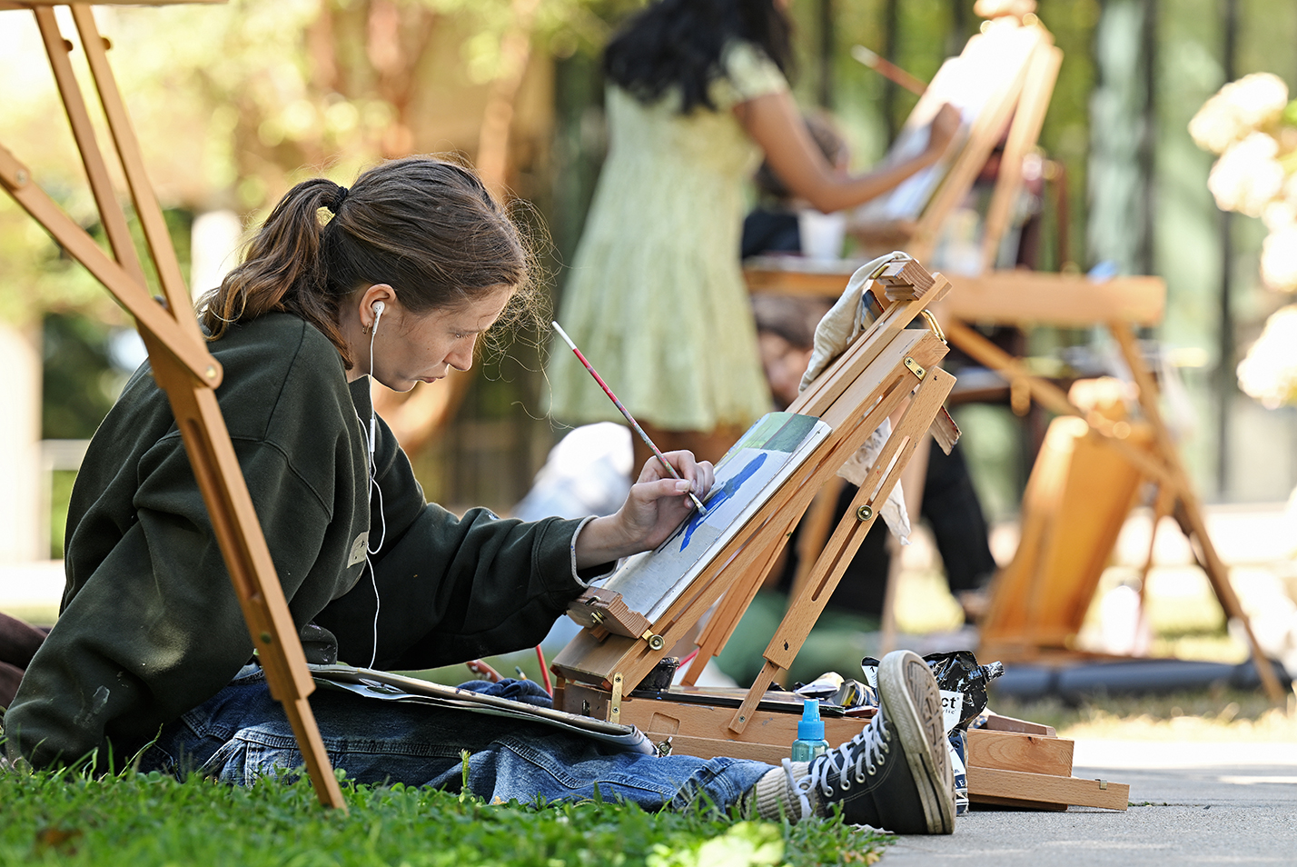 A student sits on the ground painting on an easel with other students in the background.